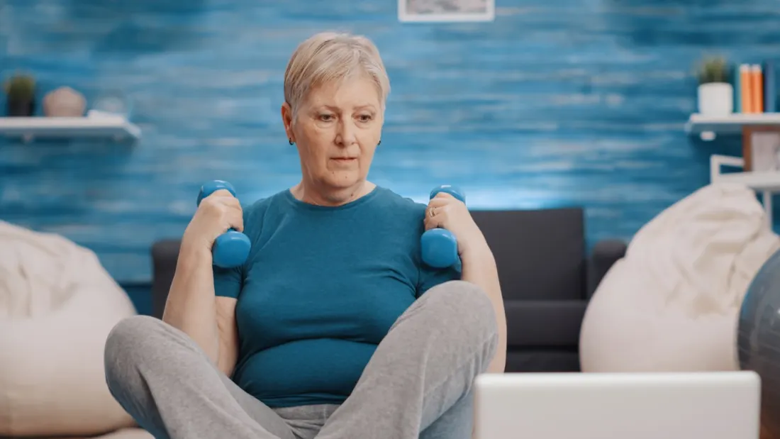 Older woman lifting dumbbells at home while following a workout video.