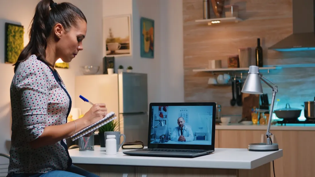 Patient taking notes during a video consultation with a doctor on a laptop.